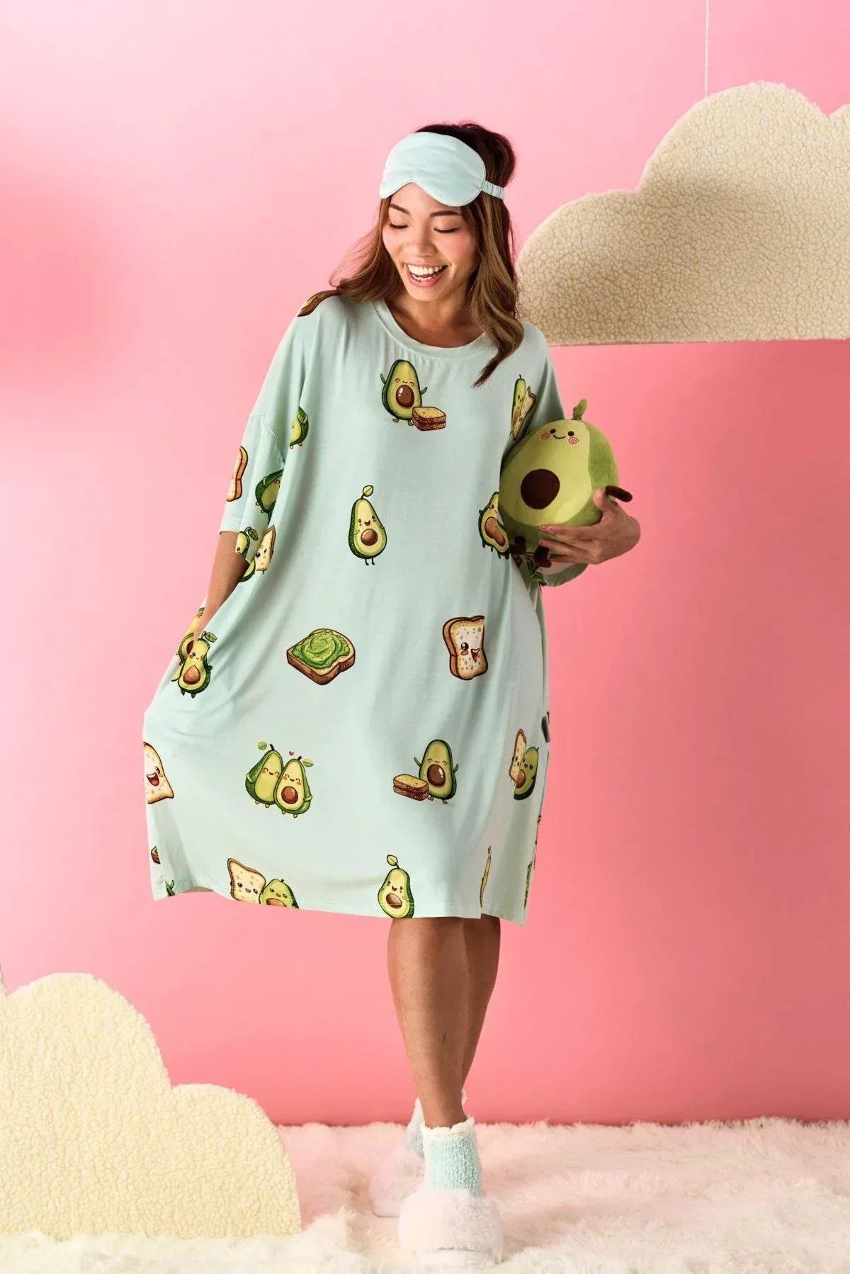 Woman wearing a light green sleep tee by MySleepTee with avocado print, holding a plush toy, against a pink background.