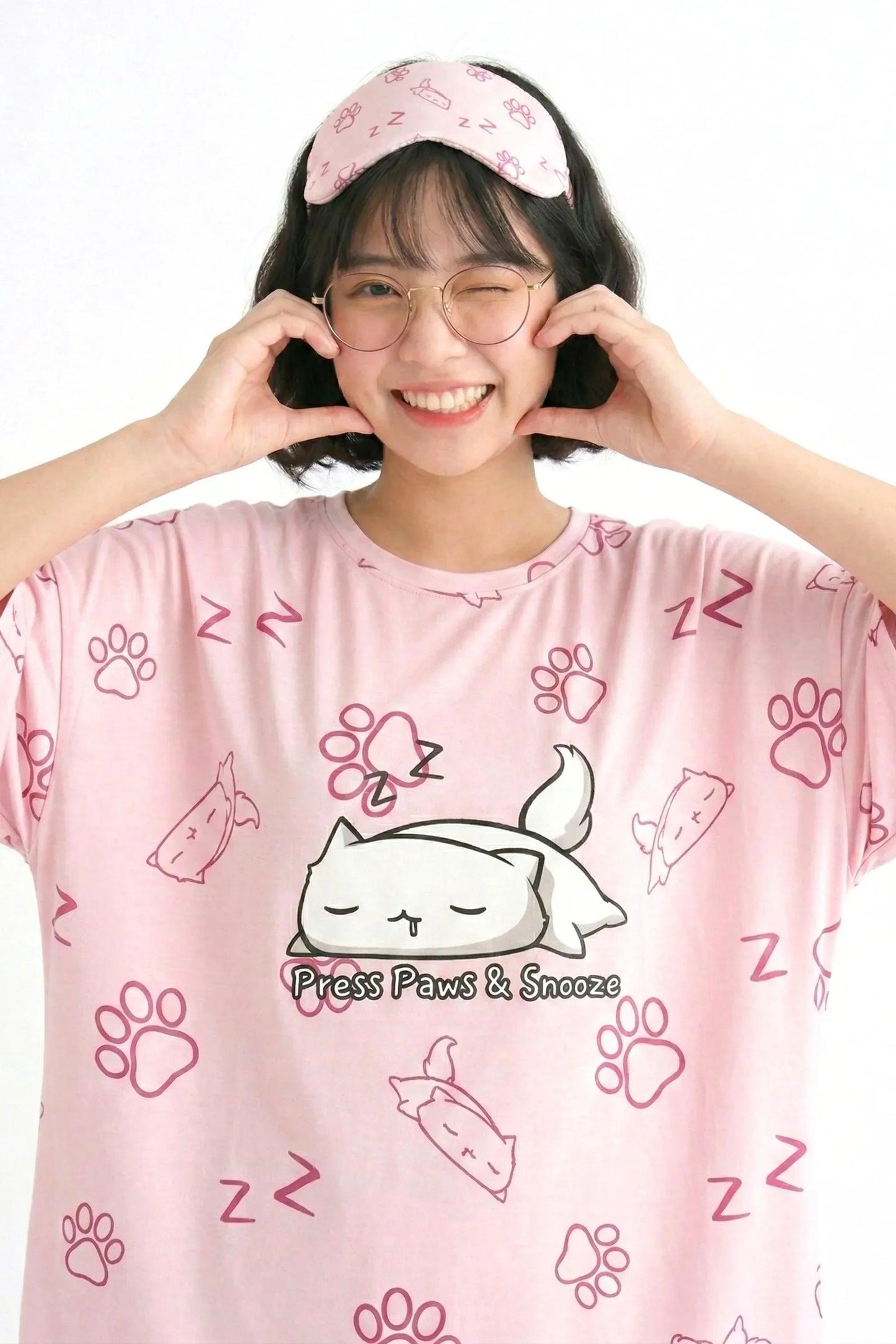 Close-up of the "Press Paws & Snooze" cat design on a pink bamboo sleep tee. The model is winking and making a heart shape with her hands.