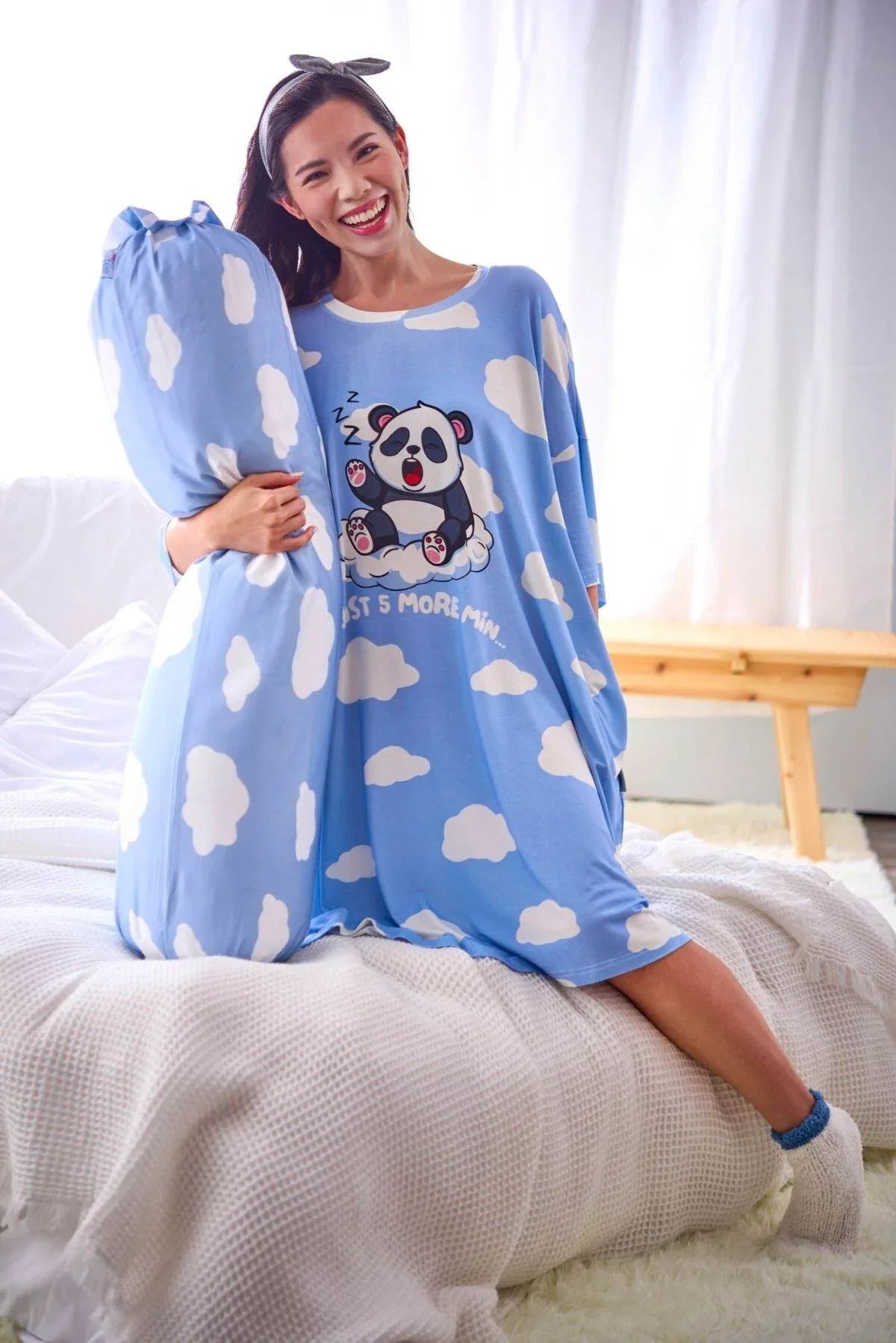 Woman wearing a blue sleep tee from MySleepTee with cloud pattern and panda design, sitting on a bed.
