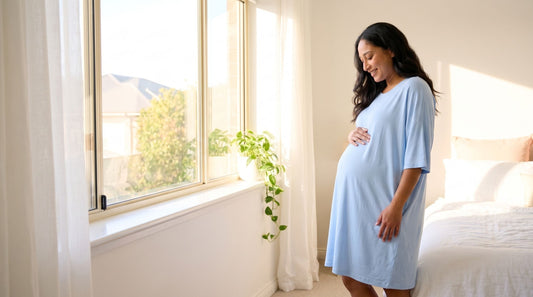 Pregnant woman in a long light blue bamboo sleep tee by a bright bedroom window