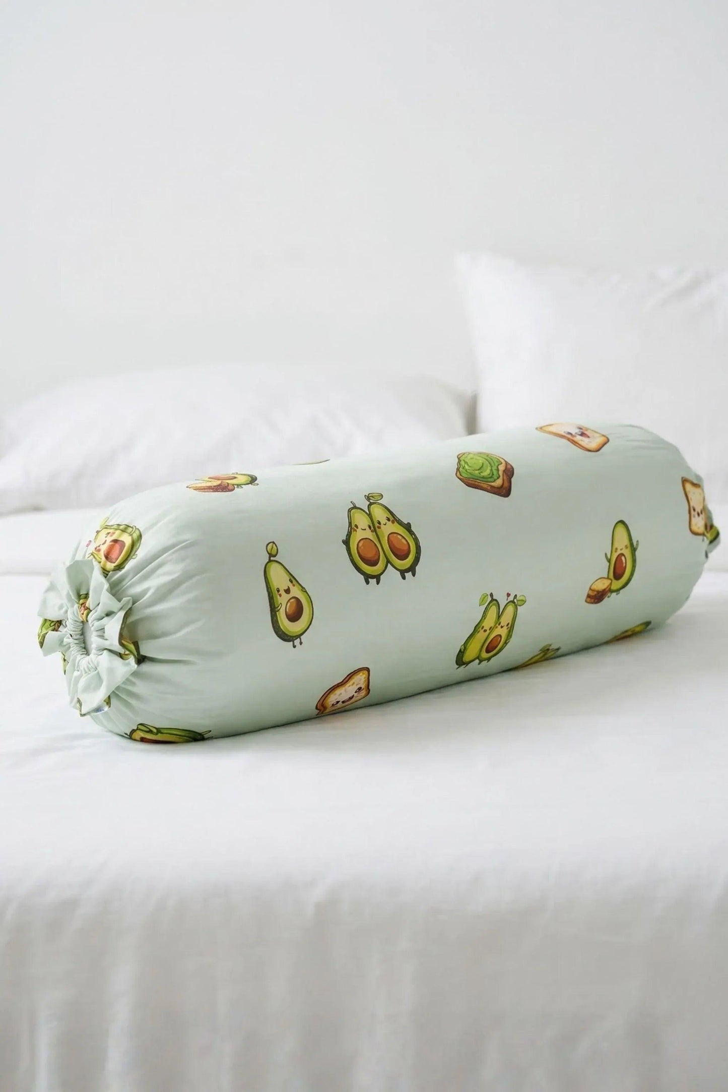 The Avo-Good Night avocado print bolster pillow lying horizontally on a crisp white bed with pillows in the background.