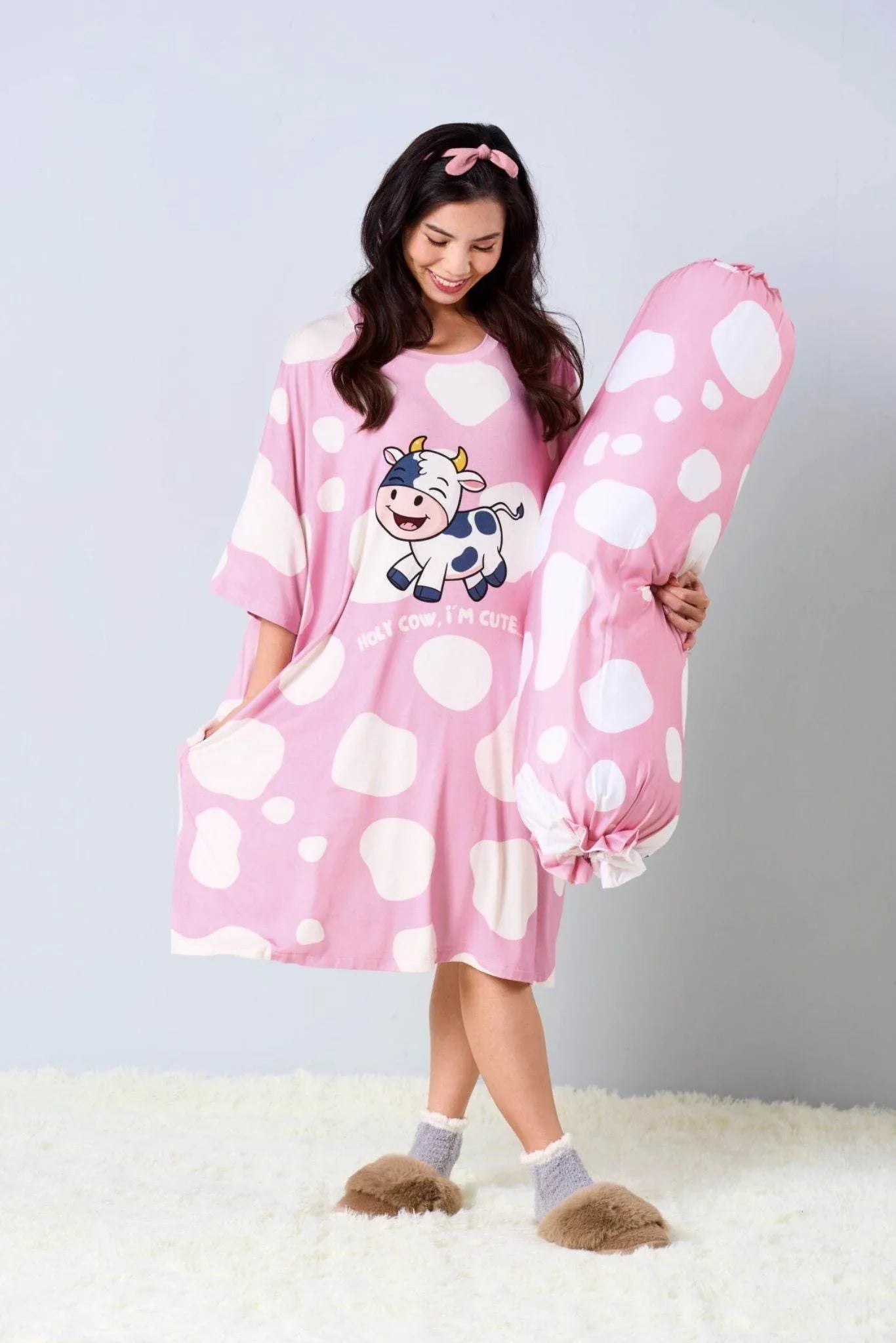 Woman wearing a oversized pink sleep tee with a cow design, holding a matching bolster.