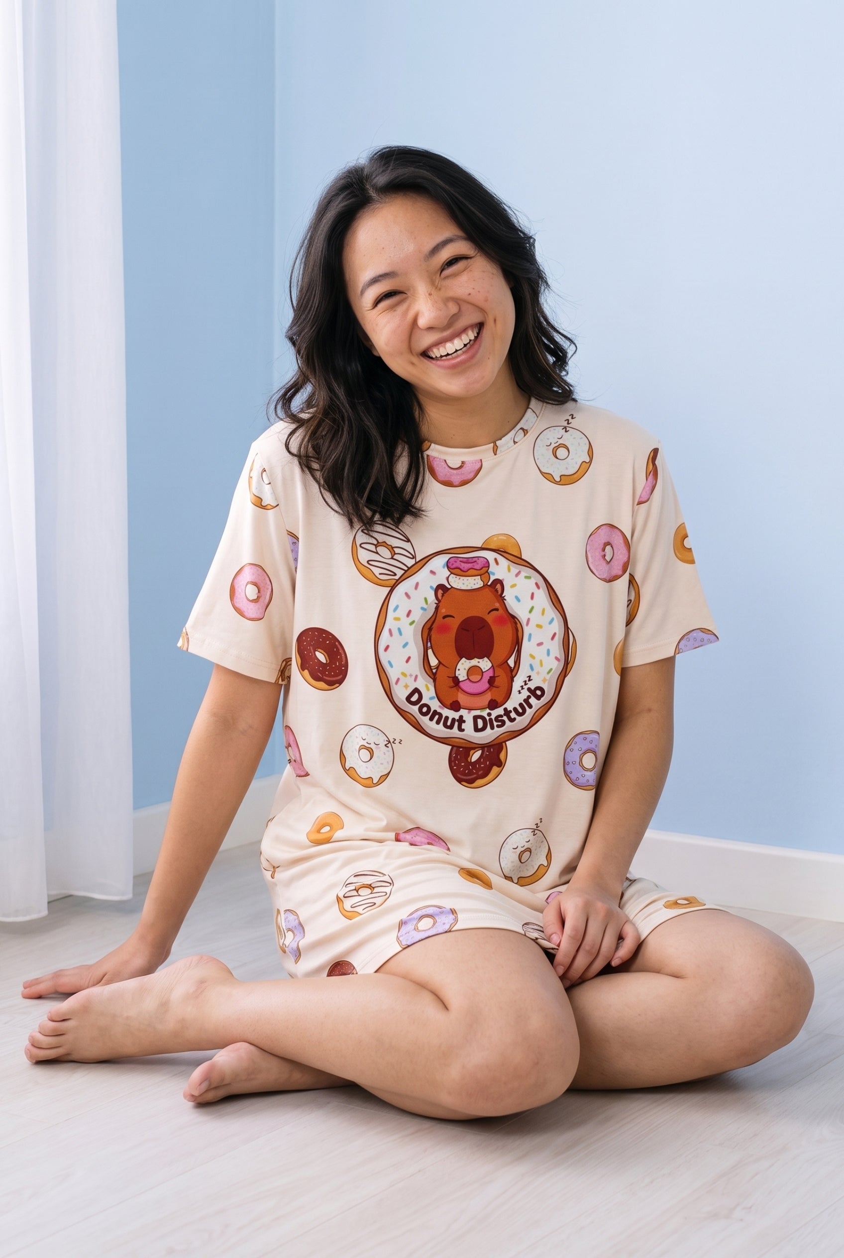 A woman sitting cross-legged on a light wood floor in a room with a light blue wall, wearing a cream-colored short-sleeve pajama shirt featuring a colorful cartoon capybara graphic and donut patterns.