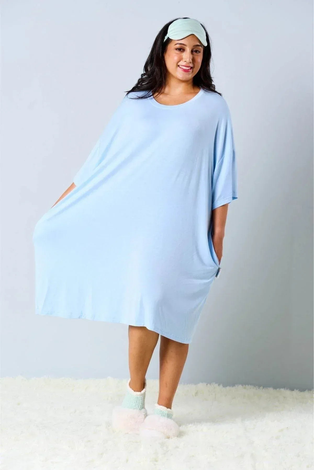 Woman wearing a light blue oversized bamboo sleep Tee by MySleepTee, white socks, and a matching sleeping mask on a plain background