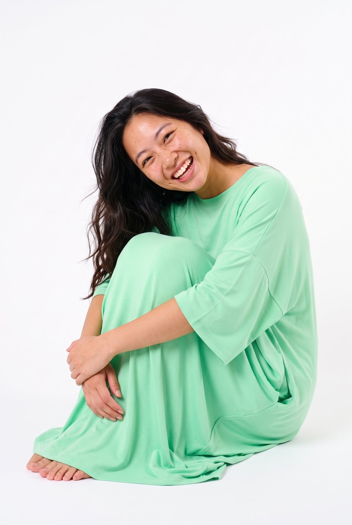 Woman laughing while sitting on the floor hugging her knees in a mint green bamboo oversized sleep tee, soft fabric draping over her legs