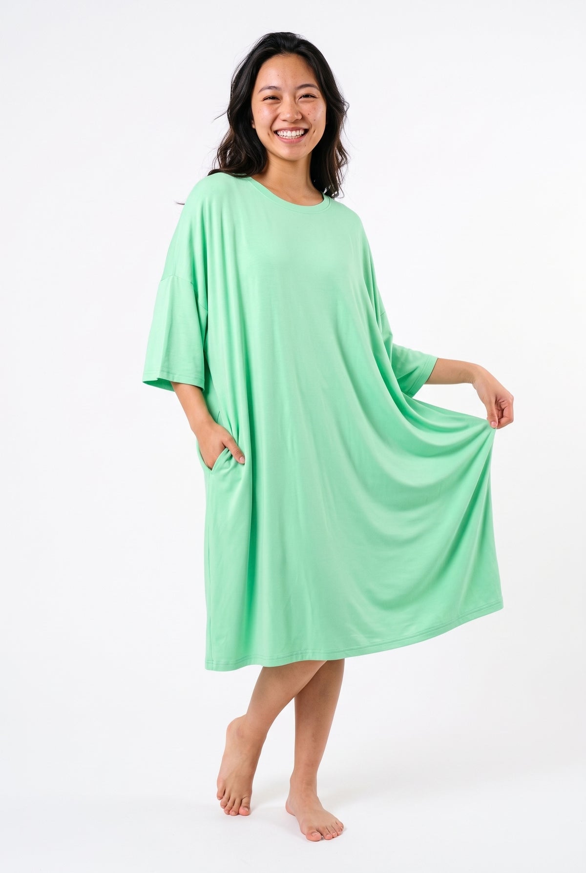 Mint green bamboo oversized sleep tee, model smiling with one hand in the hidden side pocket and the other stretching out the hem to show the roomy free-size fit