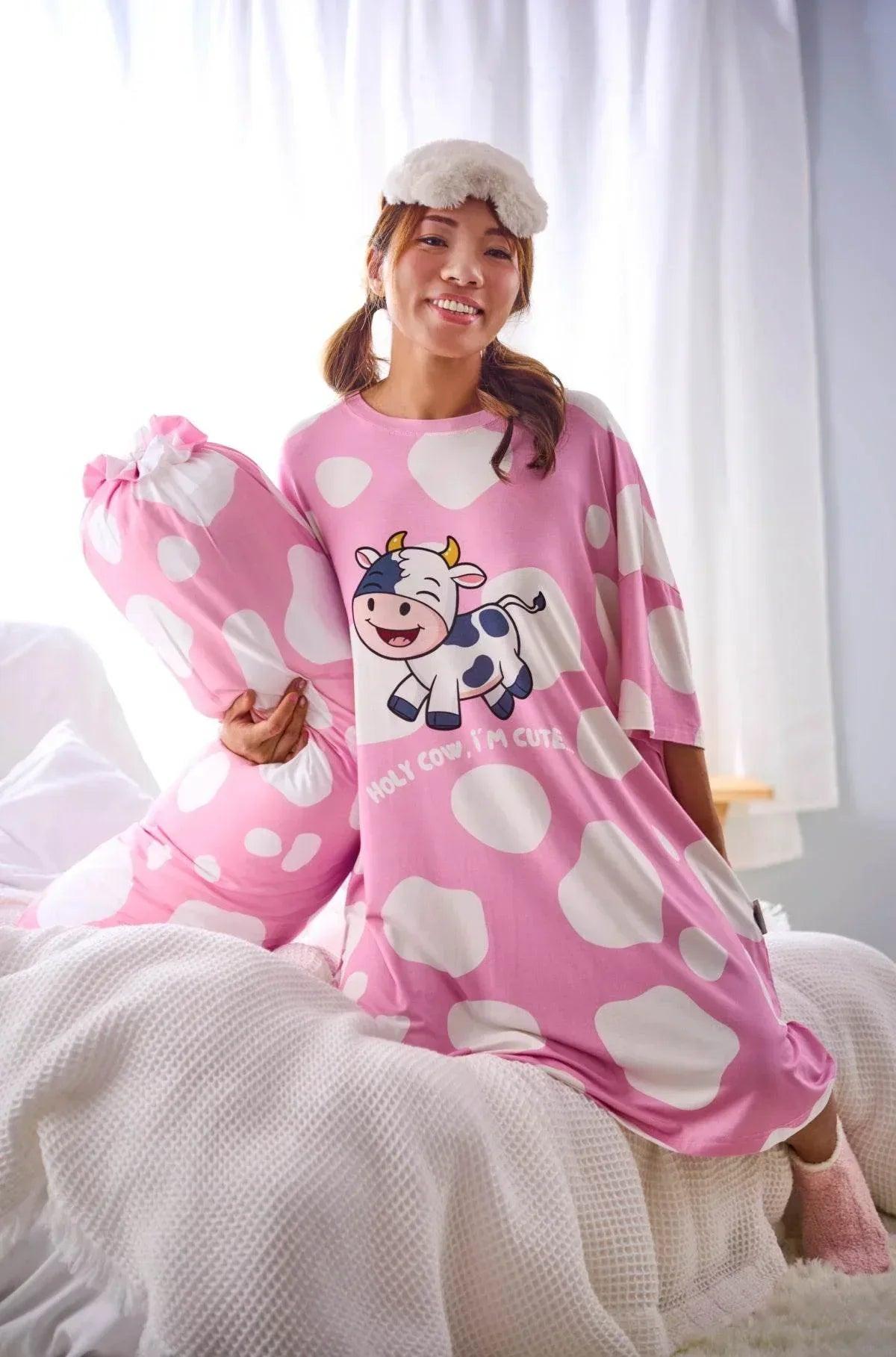 A woman in a matching pink cow print nightgown holding the Moo-nlight Bolster Cover while sitting on a cozy white bed.