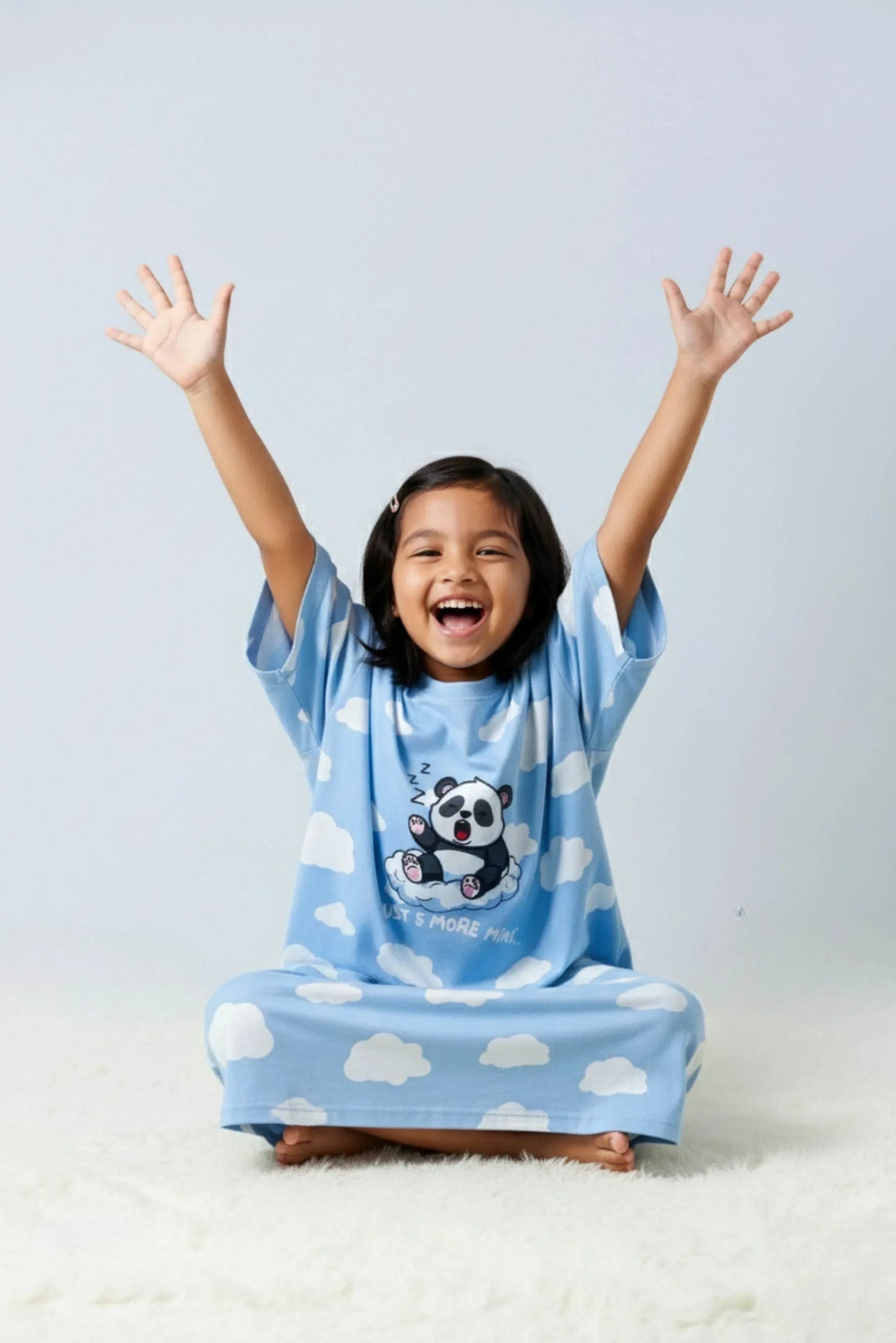 Child wearing a blue pajama set with a panda design, sitting on a white floor against a light gray background.