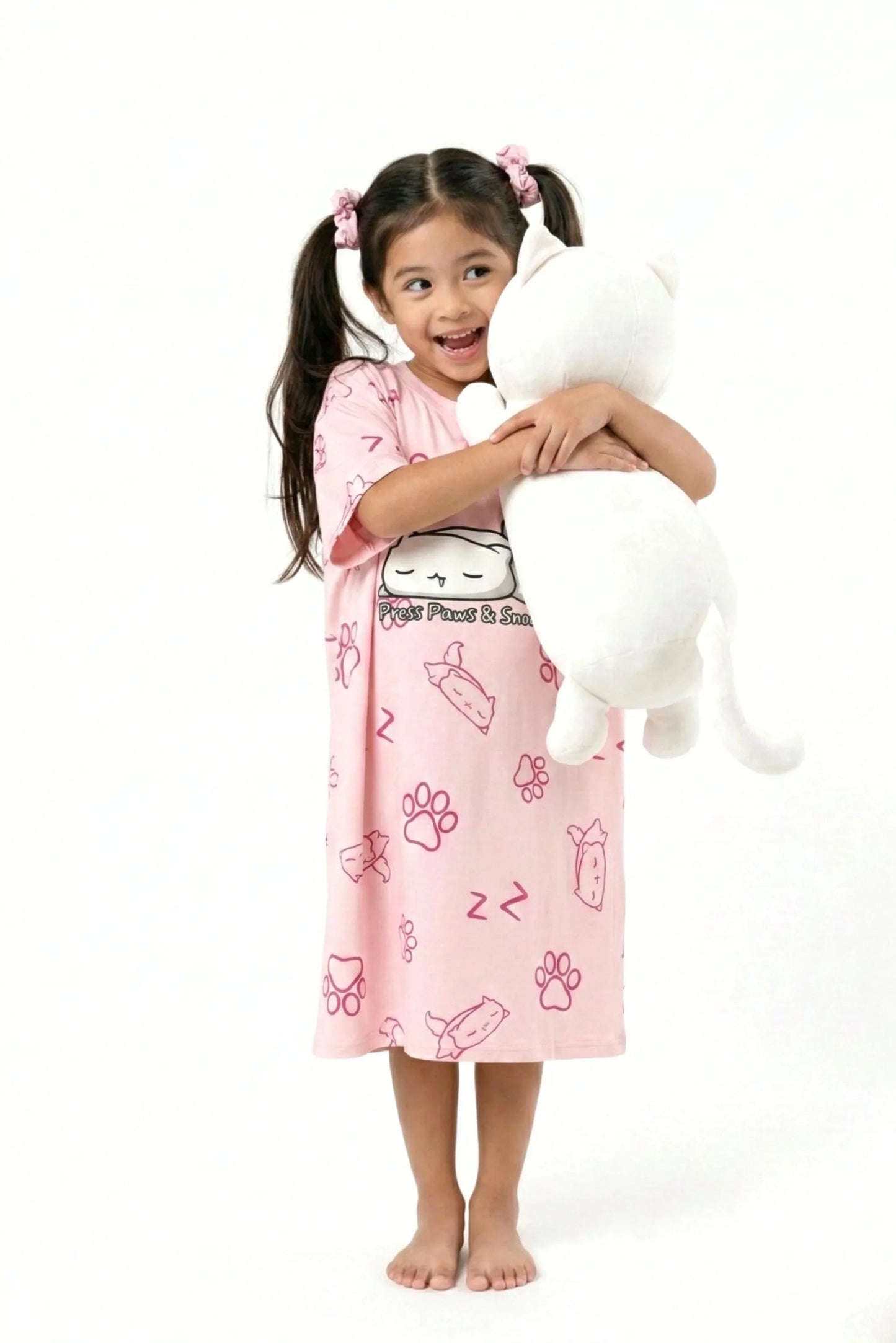 A joyful young girl hugging a large white cat plushie while wearing her MySleepTee pink bamboo oversized sleep shirt.