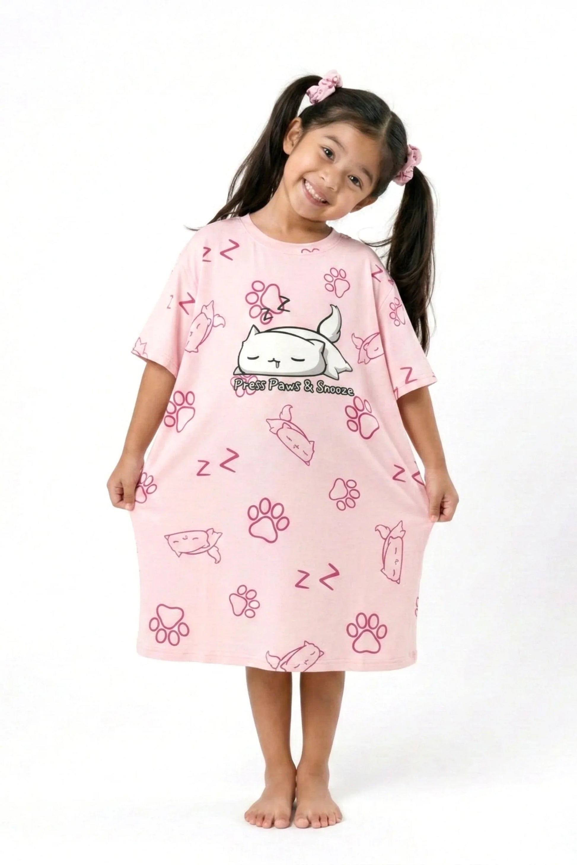 A young girl in pigtails smiling and holding the hem of her oversized MySleepTee pink bamboo sleep dress.