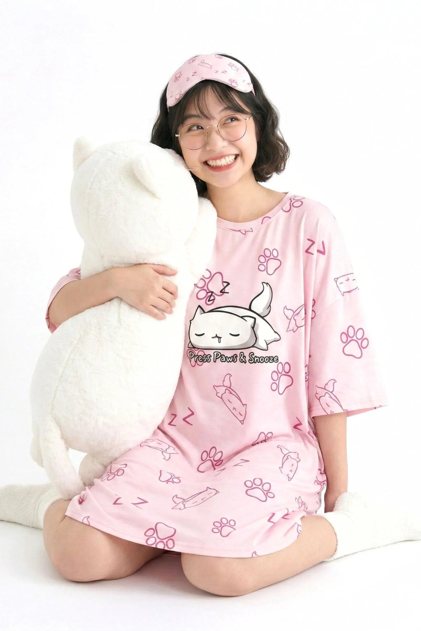 A smiling woman wearing an oversized pink MySleepTee bamboo sleep tee hugging a large white cat plushie. The tee features a "Press Paws & Snooze" cat graphic.