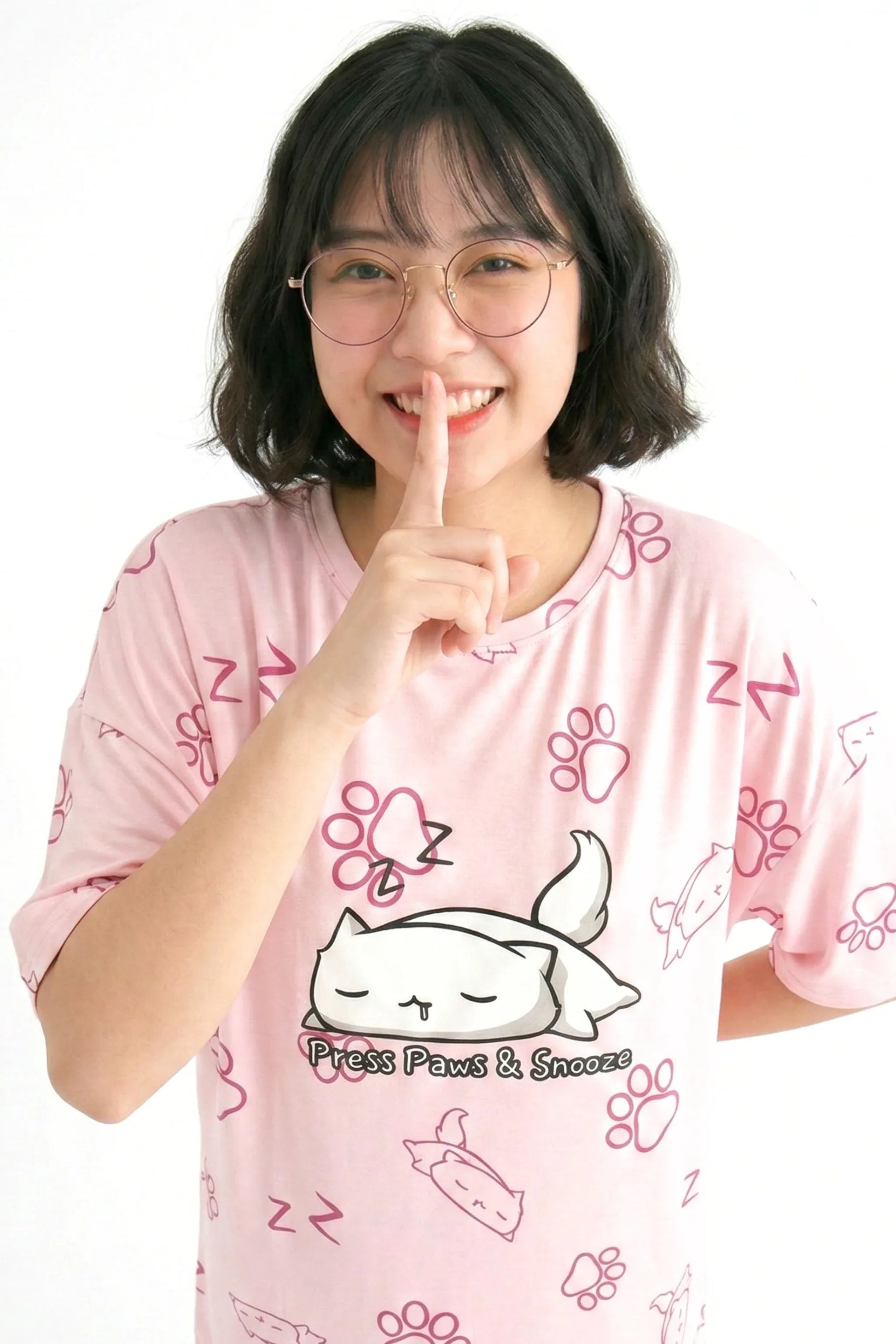 A smiling woman in a petite-length pink MySleepTee bamboo sleep tee with her finger to her lips. The shirt features a sleeping cat graphic and an all-over paw print design.