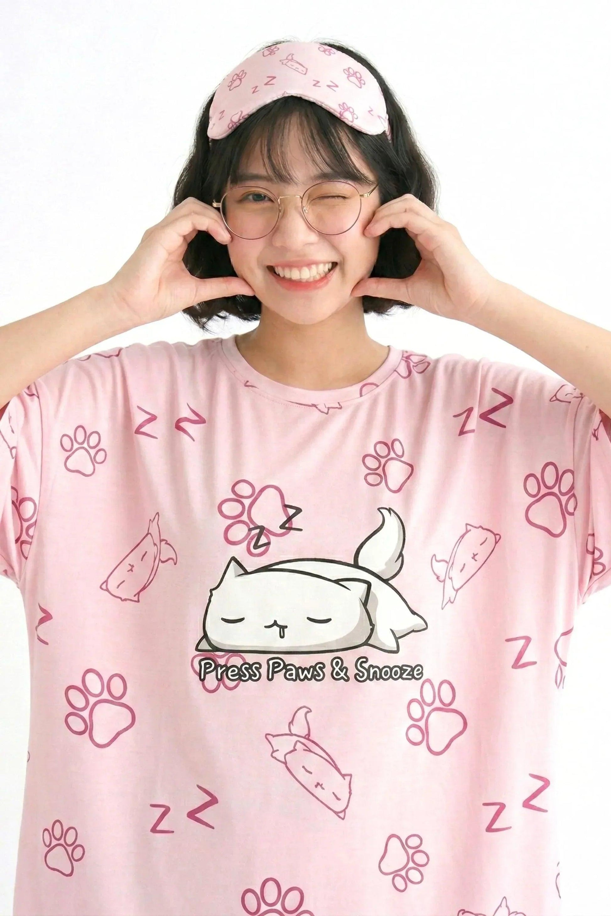 Close-up of the "Press Paws & Snooze" cat design on a pink bamboo sleep tee. The model is winking and making a heart shape with her hands.