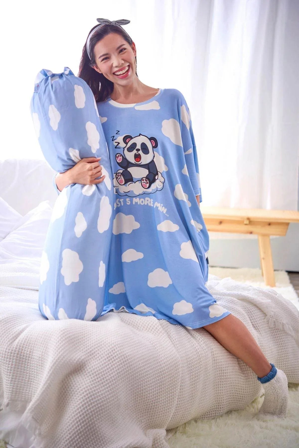 Woman wearing a blue sleep tee from MySleepTee with cloud pattern and panda design, sitting on a bed.