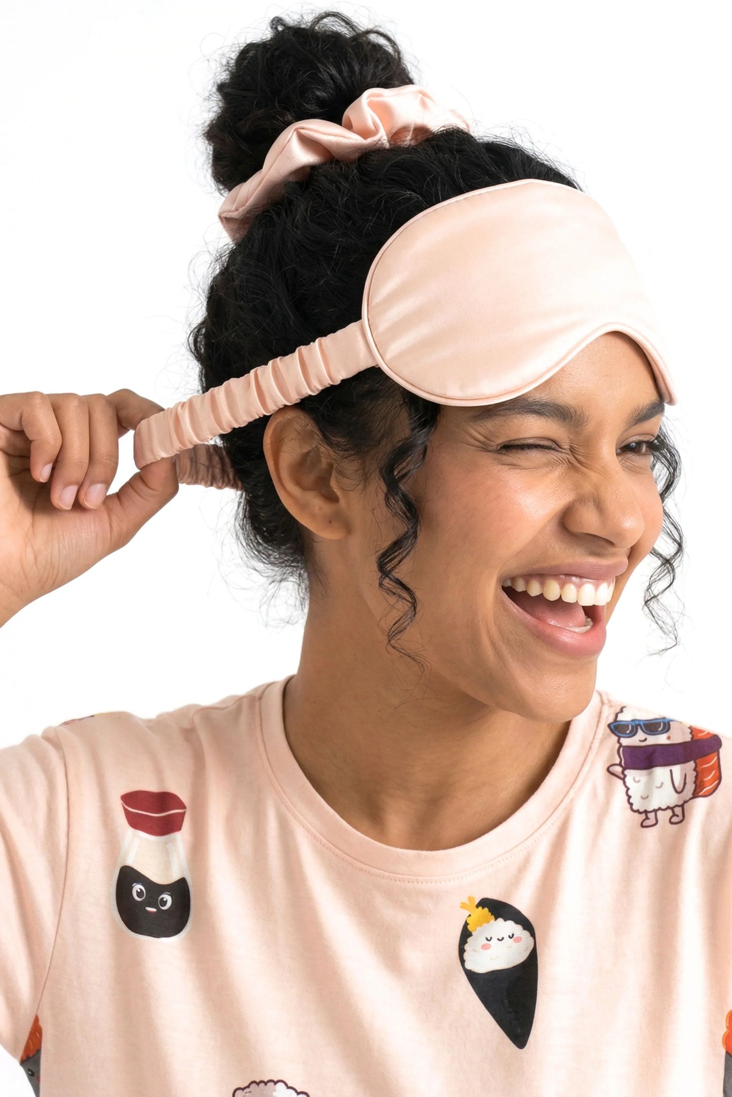 Woman laughing while pulling elastic strap of salmon pink bamboo sleep mask with matching scrunchie and Soy Cozy sleep tee