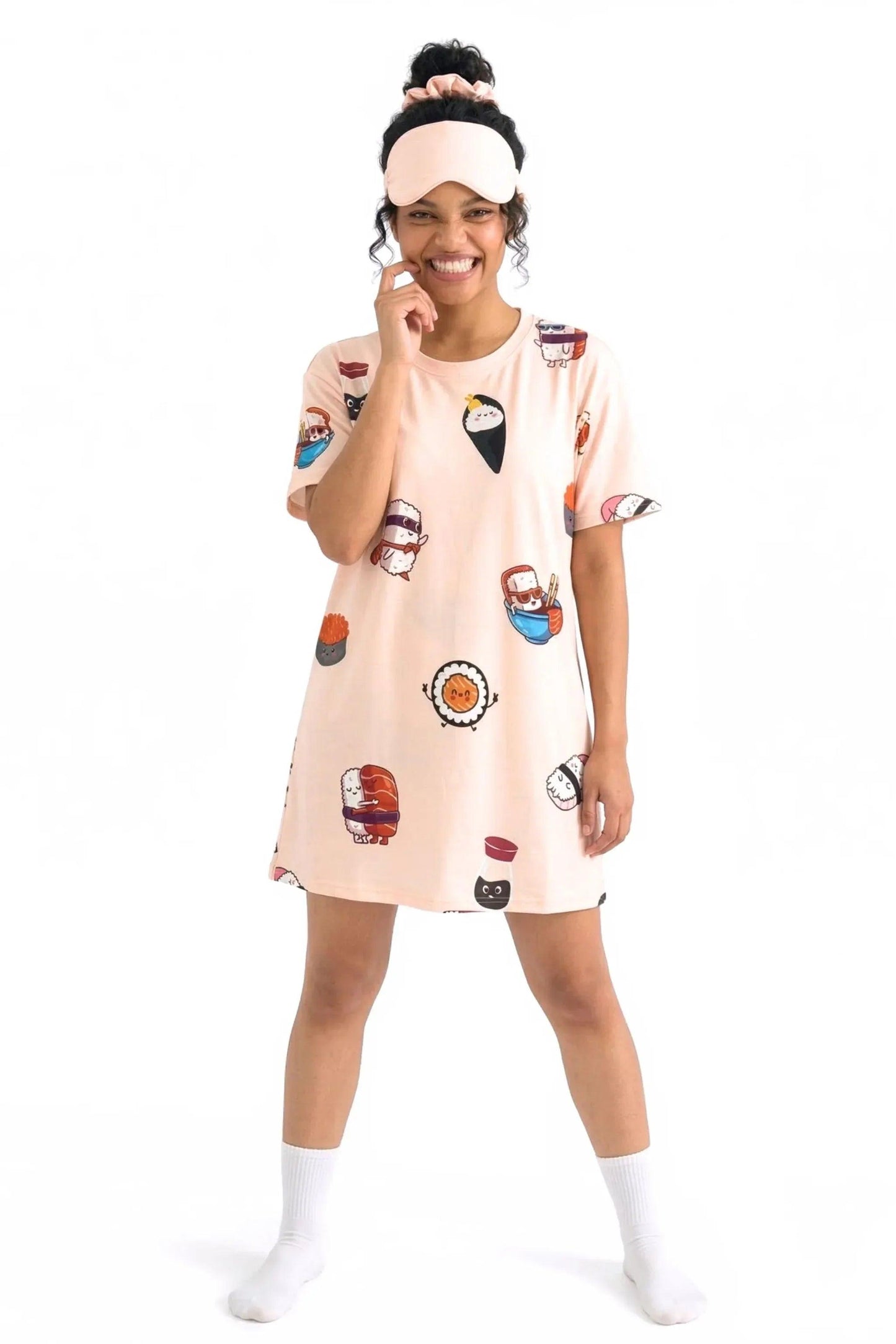 A full-body shot of a happy woman wearing the MySleepTee™ Soy Cozy Petite bamboo t-shirt dress with an adorable sushi pattern. She has a matching sleep mask on her forehead and is resting her hand near her smiling face.