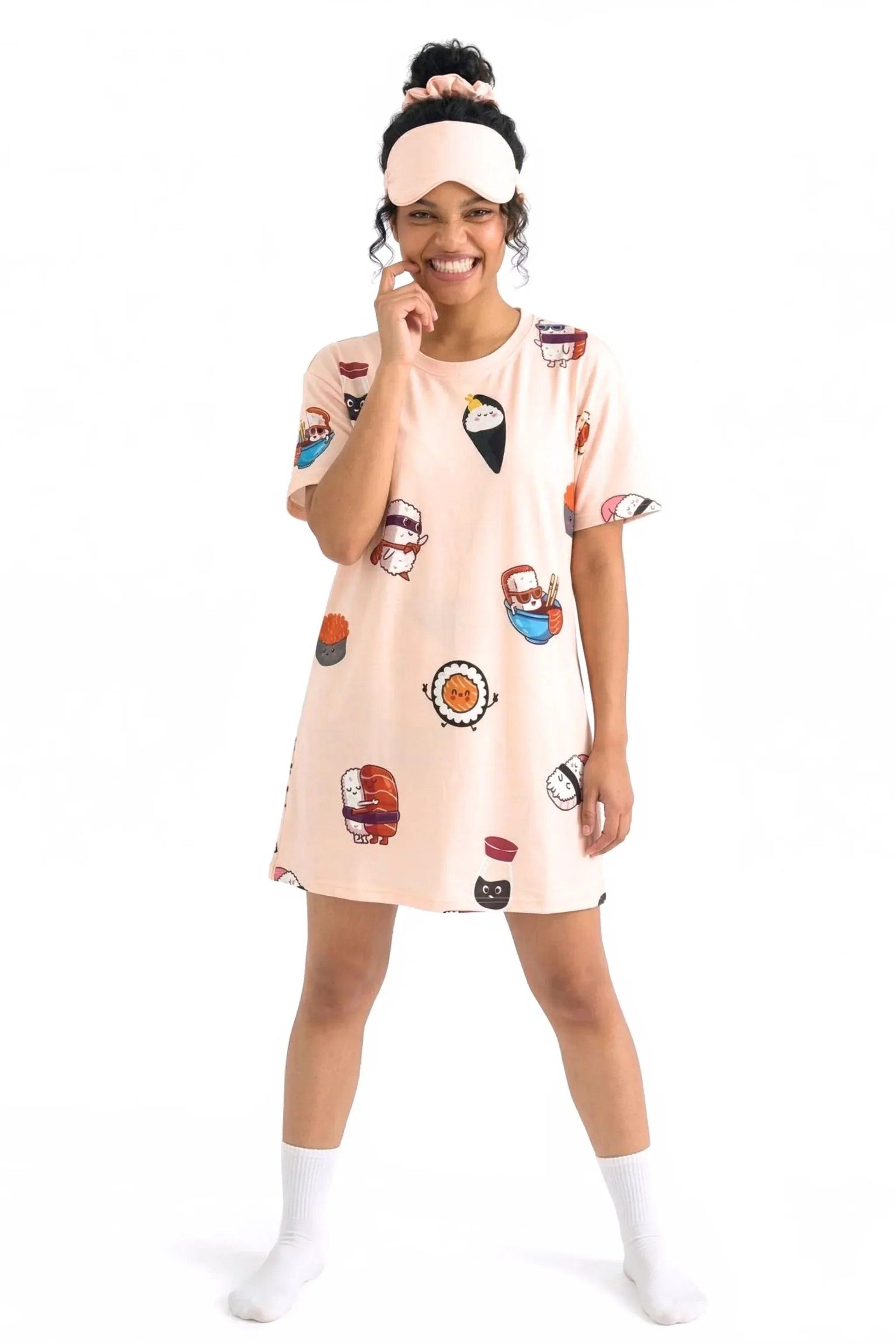 A full-body shot of a happy woman wearing the MySleepTee™ Soy Cozy Petite bamboo t-shirt dress with an adorable sushi pattern. She has a matching sleep mask on her forehead and is resting her hand near her smiling face.