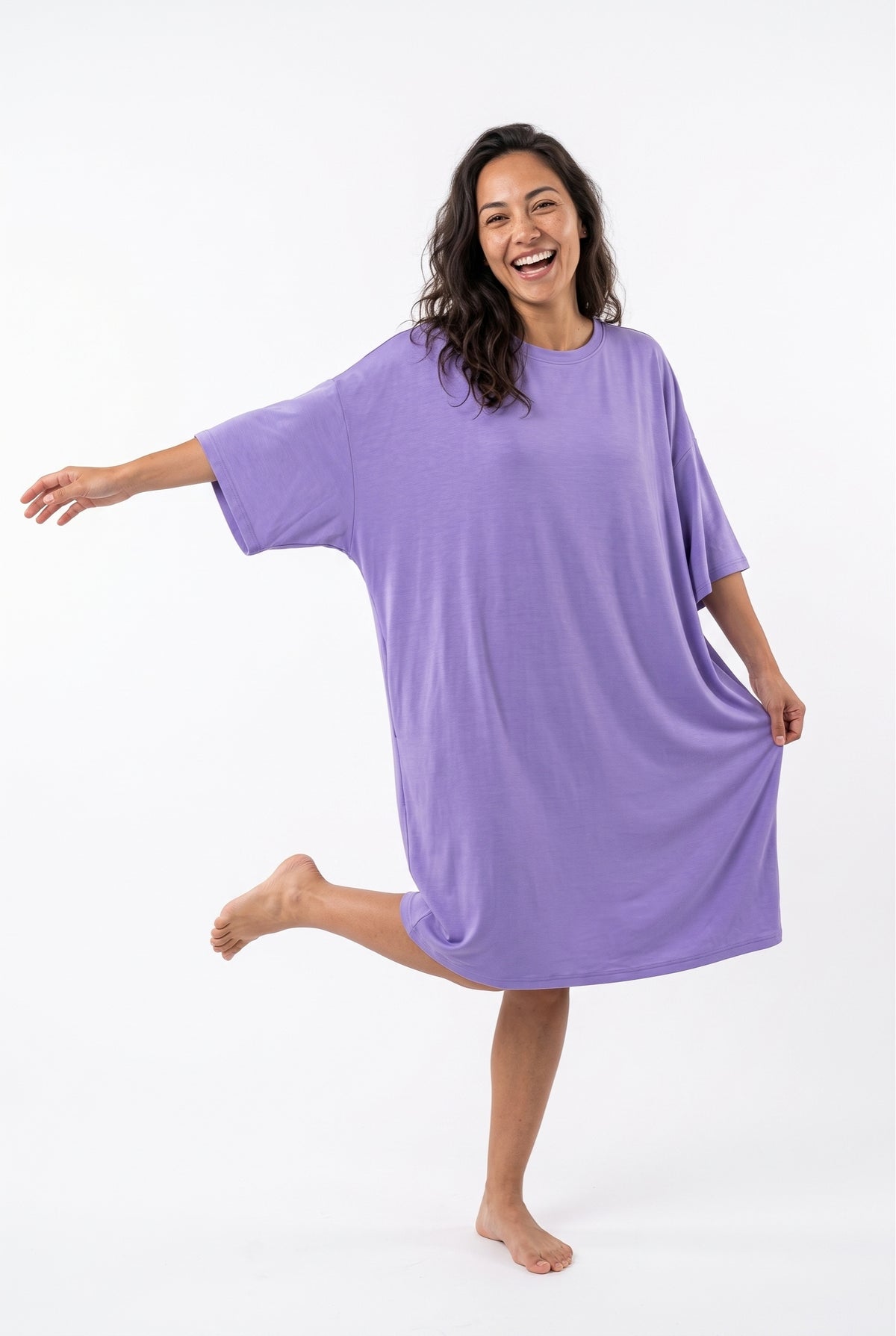 Woman laughing and kicking up one leg in a lilac purple bamboo oversized sleep tee, holding out the hem to show the roomy free-size fit