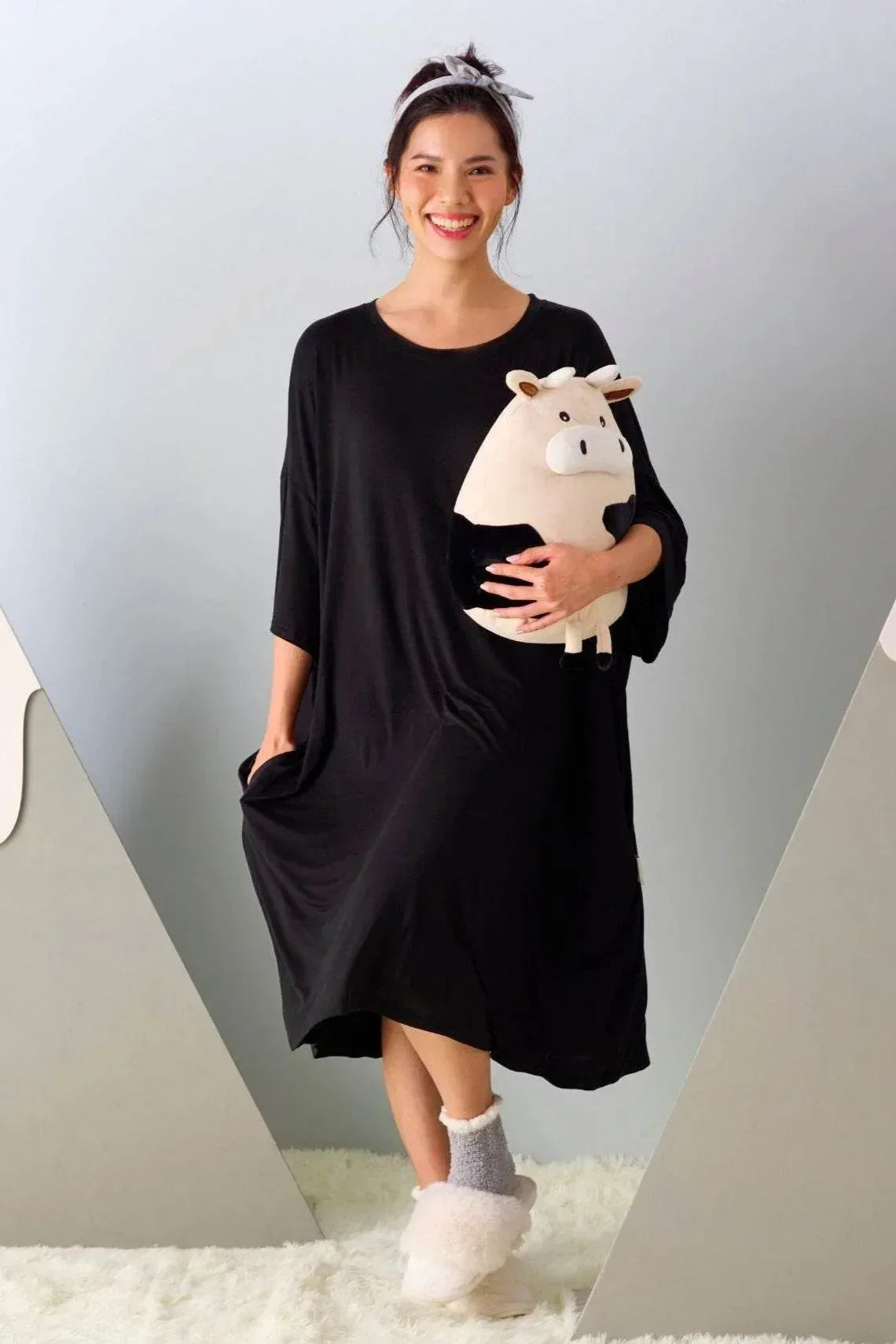 Woman in a black sleep tee holding a plush cow toy against a gray background