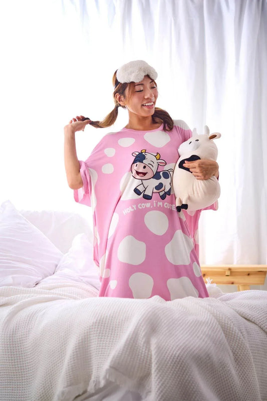 Woman in a pink sleep tee with cow design holding a plush cow toy in a bedroom.