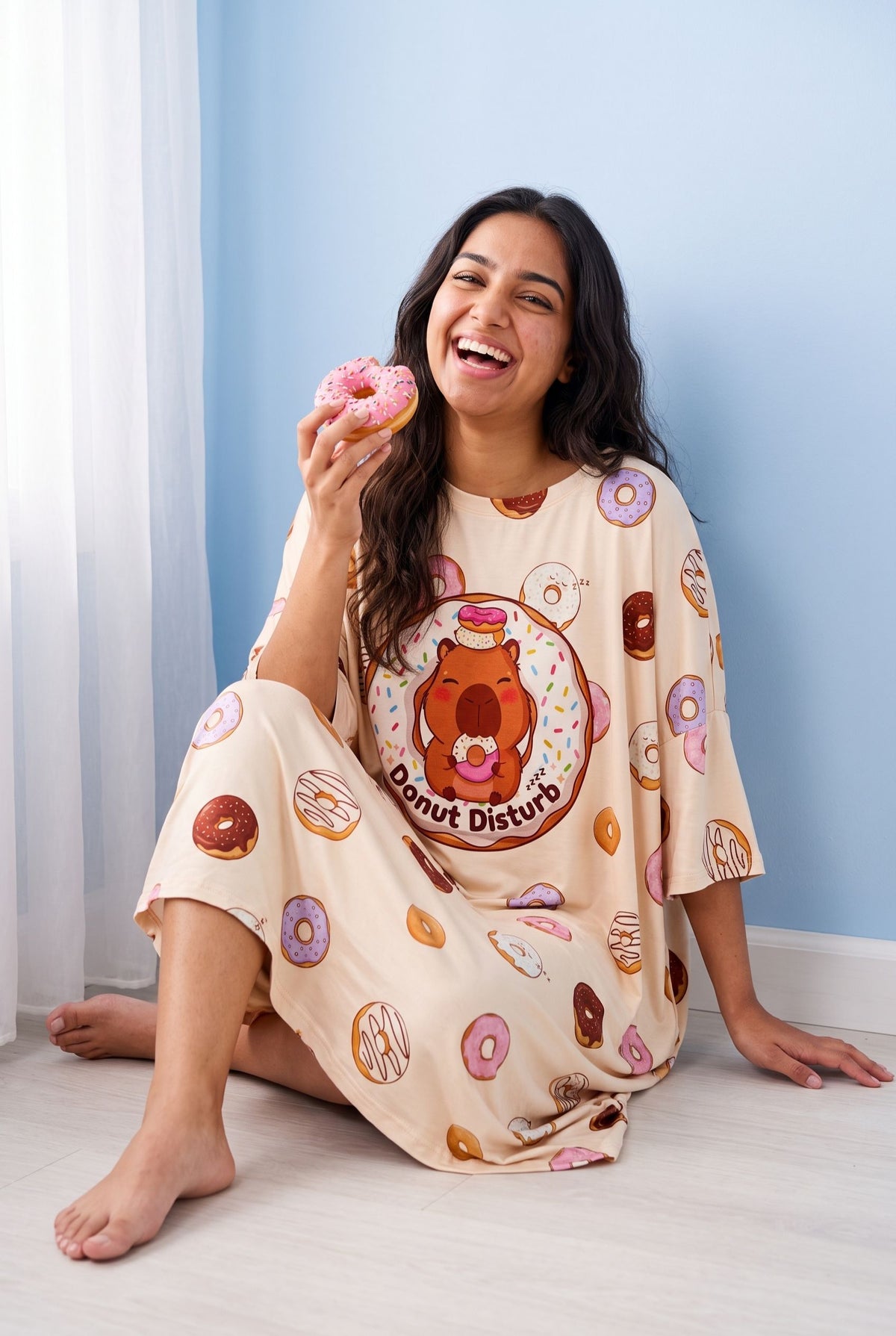 Woman sitting barefoot on a wooden floor in the Donut Disturb Original sleep tee, laughing and holding a pink sprinkled doughnut.