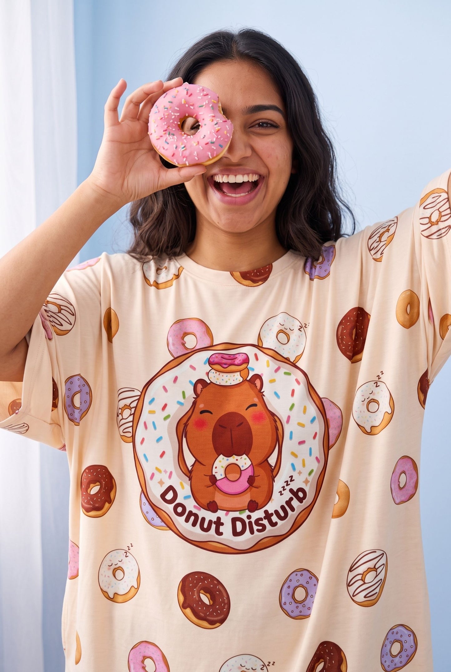 Close-up of the Donut Disturb Original bamboo sleep tee showing the sleeping capybara doughnut badge, worn by a smiling woman holding a pink doughnut.