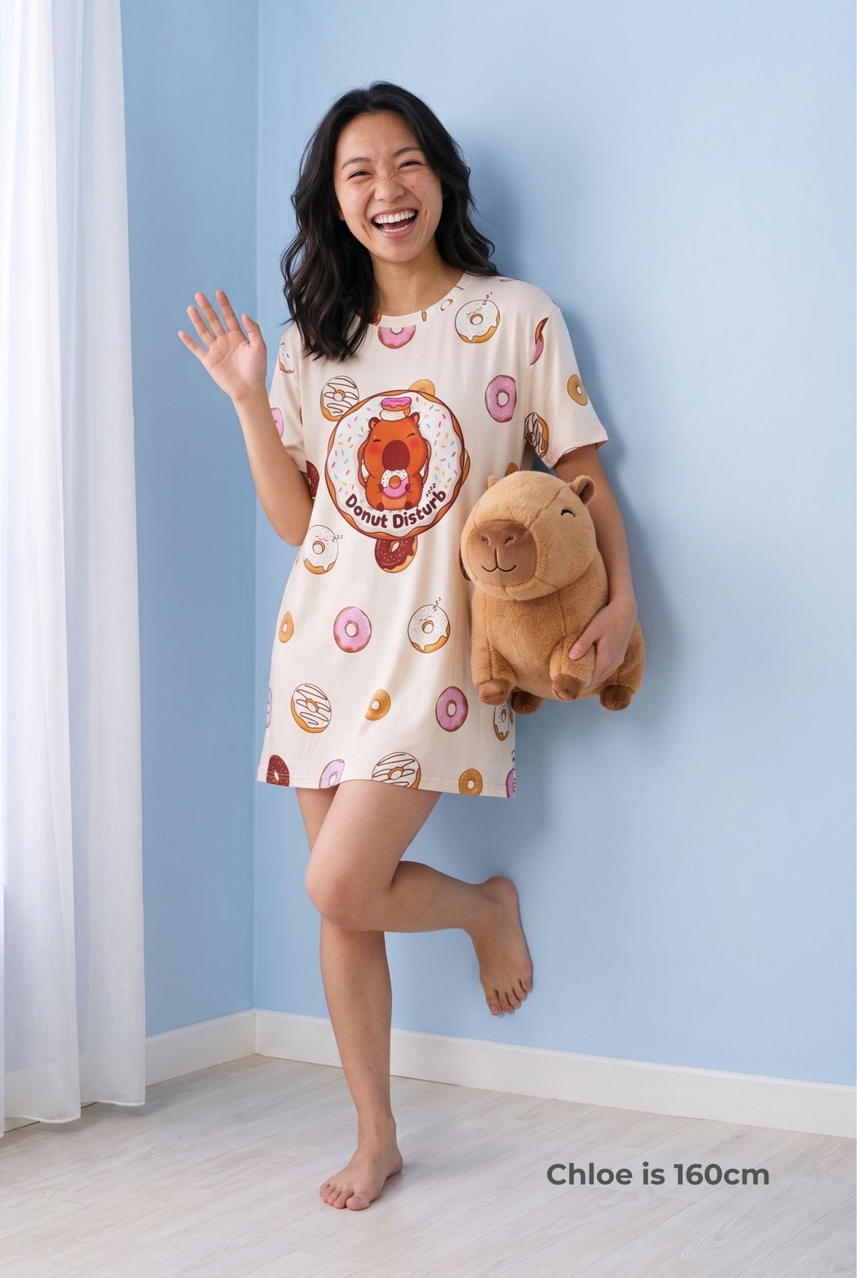 Woman in the Donut Disturb Petite bamboo sleep tee holding a capybara plush toy and waving, hem falling mid-thigh.