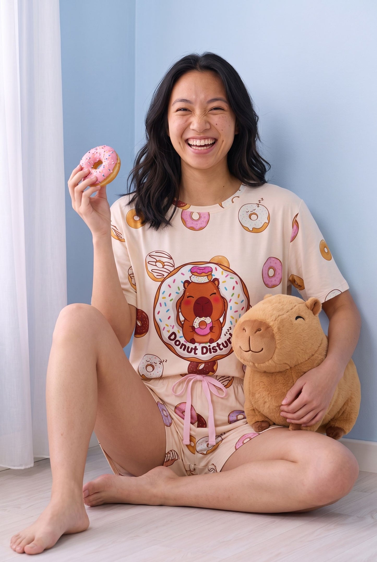 Woman sitting cross-legged on a wooden floor in the Donut Disturb tee and shorts set, laughing while holding a pink doughnut and a capybara plush toy.